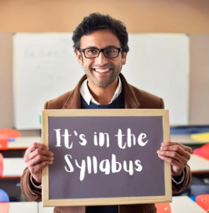 A smiling professor with glasses and dark curly hair is standing in a classroom, holding a framed sign that reads, "It's in the Syllabus." He is wearing a brown coat over a sweater and collared shirt. The classroom is brightly lit with colorful chairs and a whiteboard in the background.