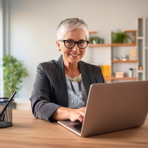 A middle-aged woman with short gray hair and glasses is sitting at a desk, smiling and using a laptop. She is dressed in a striped blazer and a light-colored blouse. The background features a modern office with shelves containing books and plants, creating a friendly and efficient atmosphere. A pencil holder is also visible on the desk.