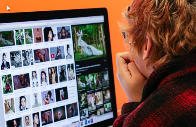 A student with short, curly hair and glasses is sitting in front of a computer screen, intently looking at various images displayed on the monitor. The screen shows a collection of photographs, some of which appear to be fashion or nature-related. The background is orange, adding a vibrant contrast to the scene. The student is wearing a red and black flannel shirt and appears focused on their task.