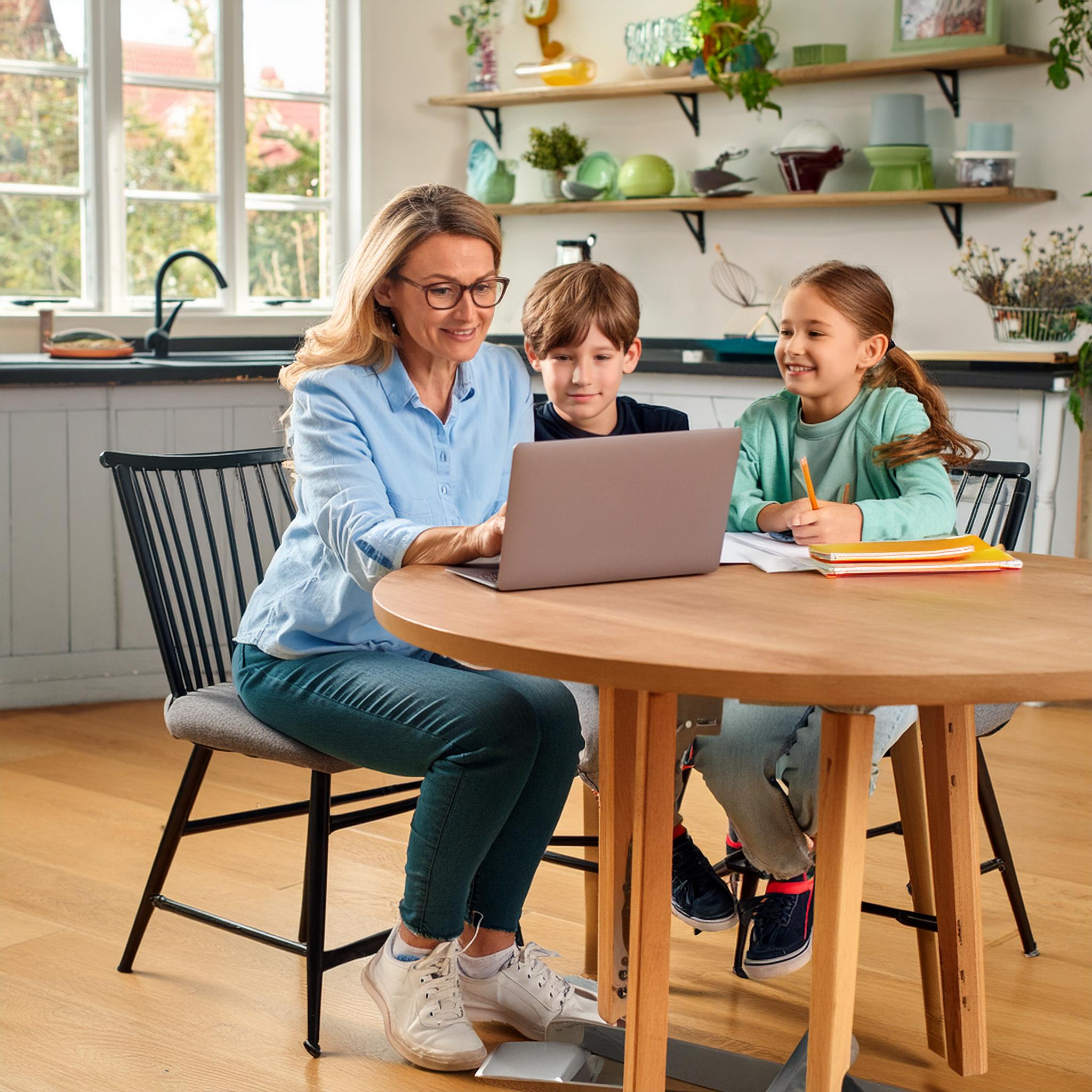 A woman, who appears to be a nontraditional student, is sitting at a kitchen table with her two young children. She is using a laptop, while the children, one boy and one girl, are attentively watching and engaged with her work. The kitchen is bright and modern, with shelves displaying various dishes and plants in the background.