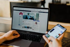 Person looking at their smartphone with a social media page on their laptop in the background.